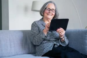 Eine lächelnde Seniorin sitzt auf einem Sofa und hält ein Tablet in der Hand, auf das sie schaut.
