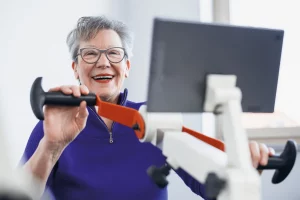 Eine lachende Seniorin mit Brille und lila Pullover bewegt mit den Armen ein Übungsgerät des VitaTables und schaut auf ein Tablet.